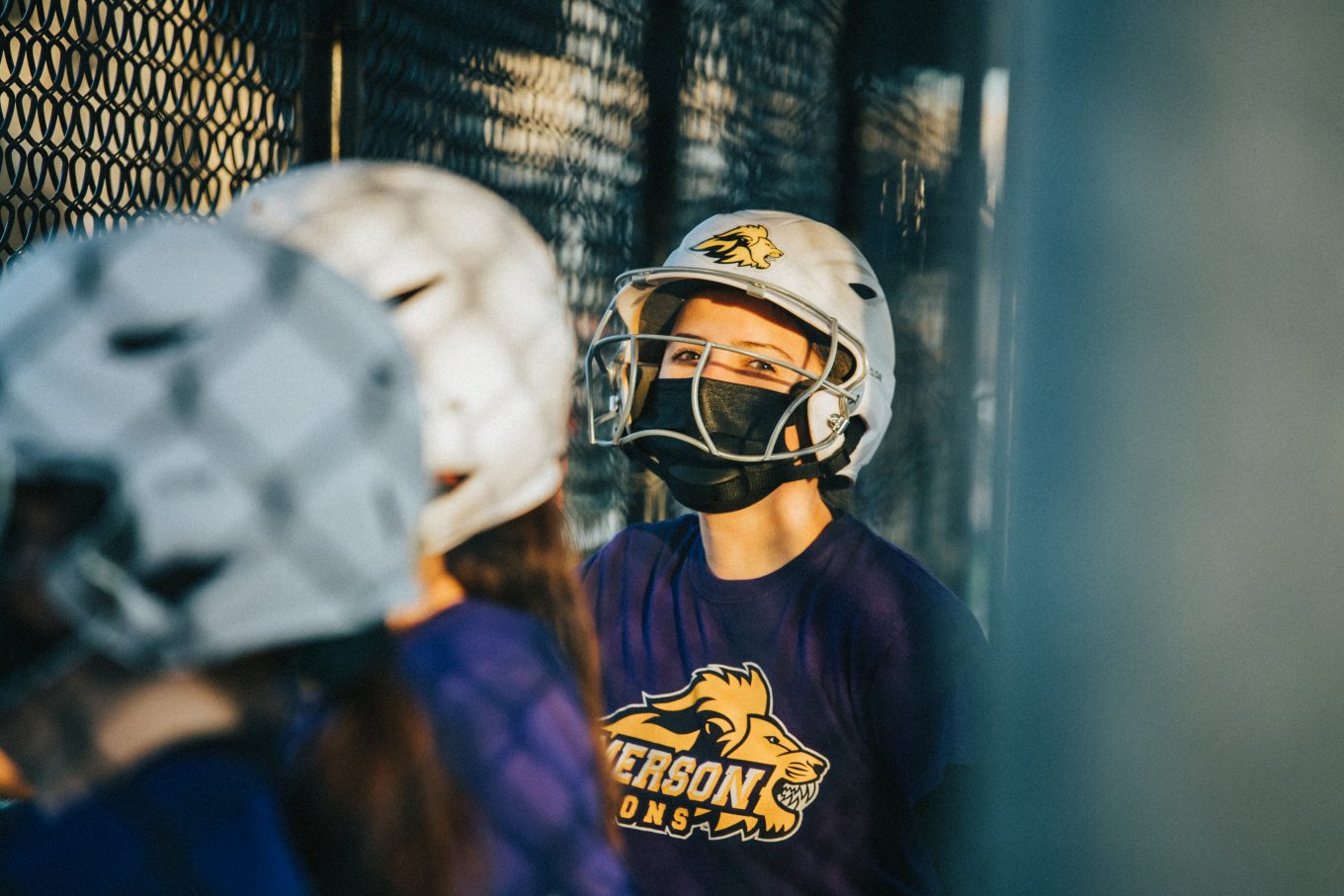 Softball player wearing a helmet and an Emerson Lions t-shirt standing by a fence during softball practice