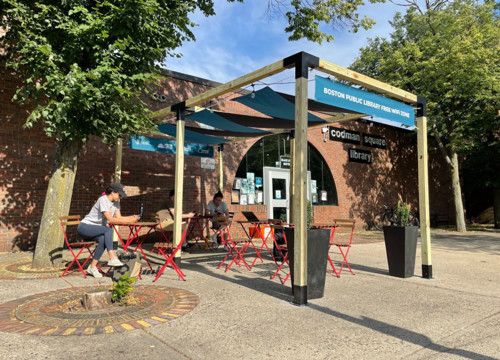 Codman Square pop-up library with sign reading "Boston Public Library Free Wifi Zone" with people sitting outside at the tables reading