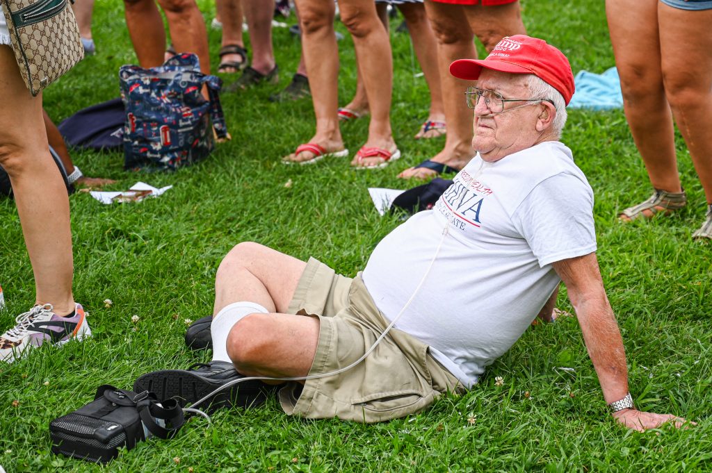 An older man sitting cross-legged in the grass wearing a red hat